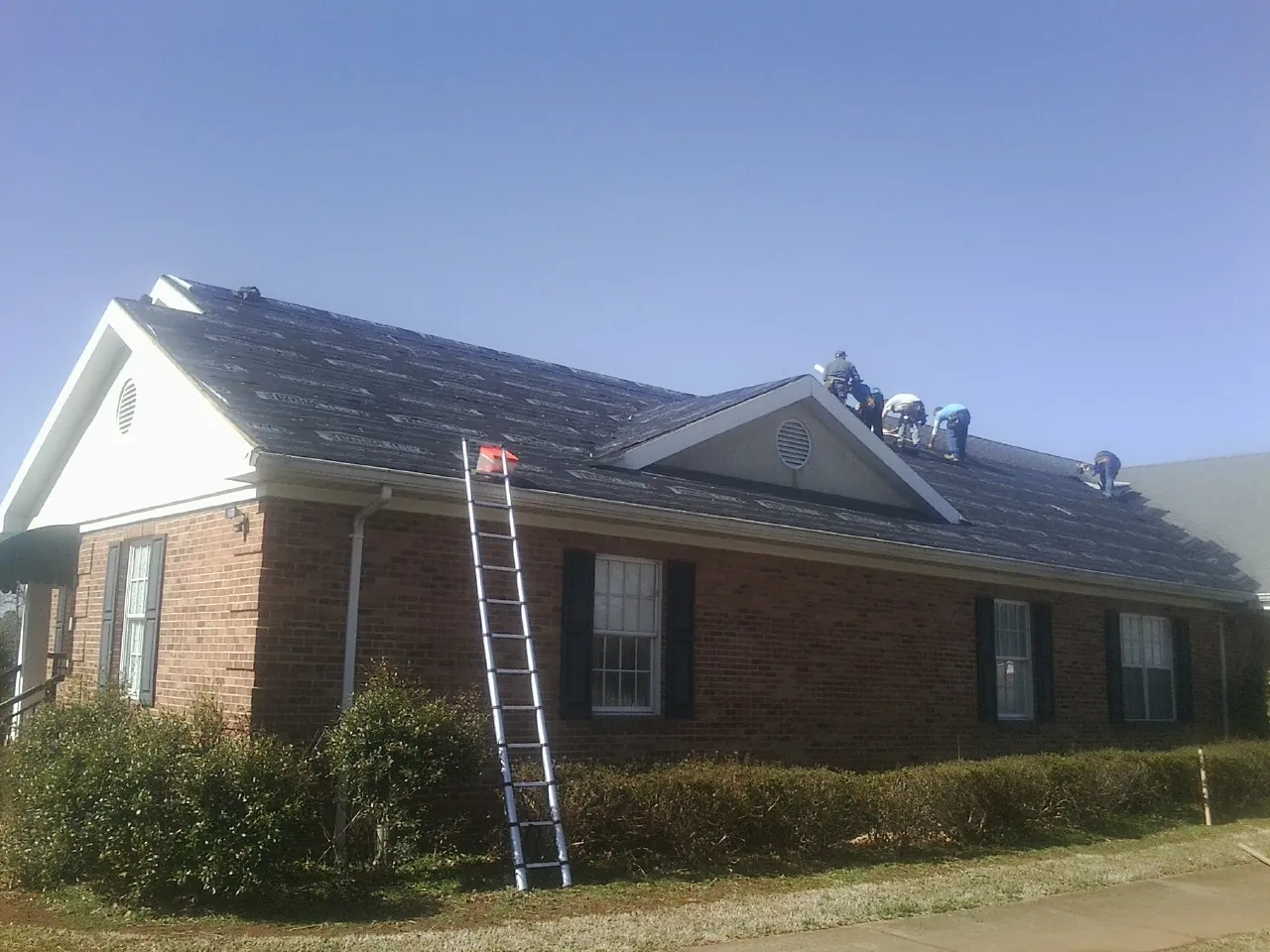 Crew tearing shingles off a brick ranch roof with a ladder set — Emergency Roof Repair work by our crew in Maple Grove