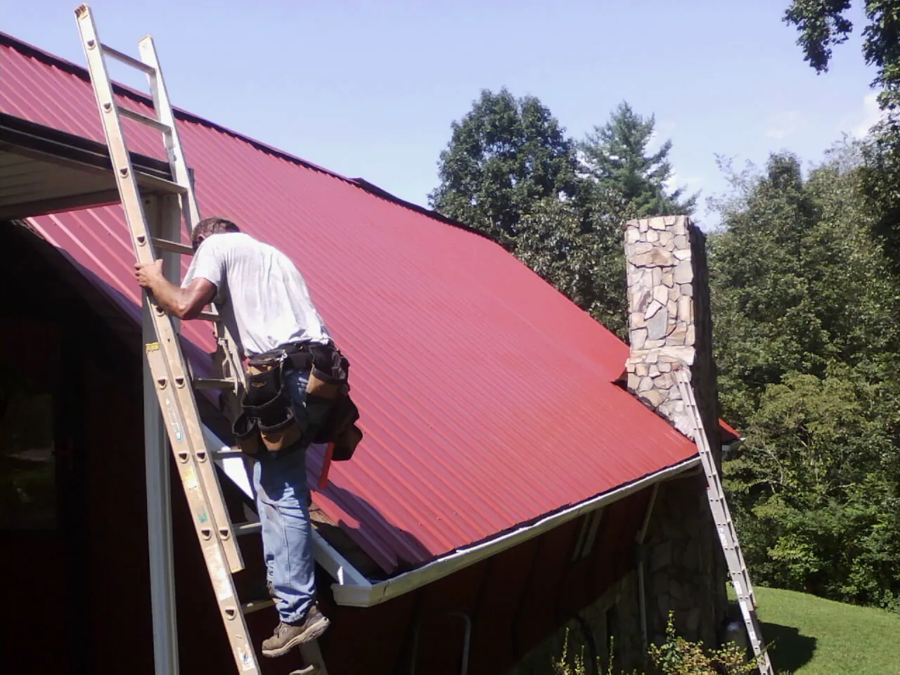 Roofer on a ladder beside a red metal roof and stone chimney — Soffit & Fascia Repair by Roofing Brothers in Maple Grove